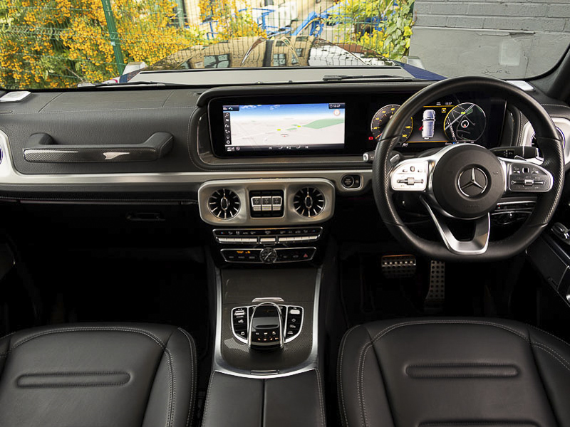 Inside a Mercedes-Benz G-Class cockpit showing steering wheel, dashboard, centre console, infotainment screen and front seats 