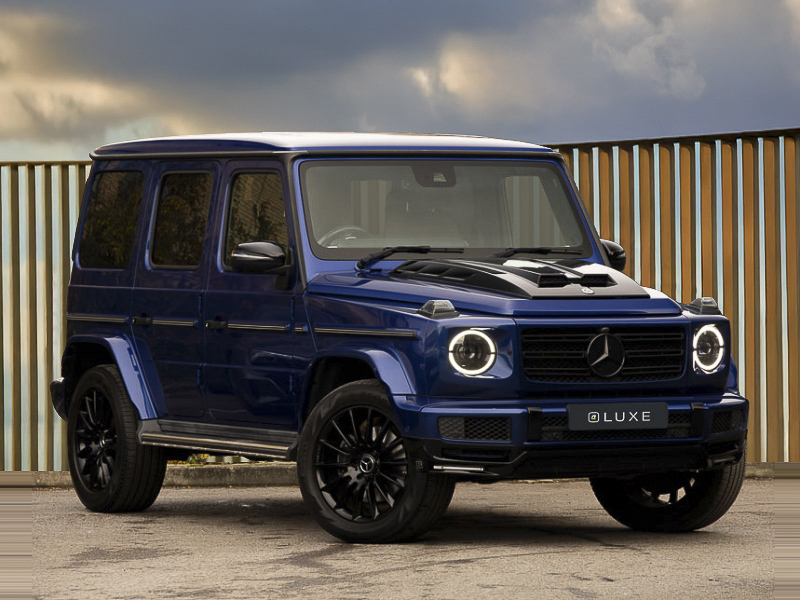 A blue Mercedes-Benz G-Class from the front parked on paving stones with a fence and cloudy sky in the background